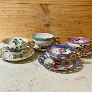 May include: Four antique teacups and saucers are arranged on a white surface. Each cup features a unique floral design and gold-colored handles. The saucers have matching patterns and gold trim, creating an elegant tea set.
