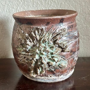 May include: A handmade, rustic ceramic pot with a textured surface. The pot is primarily brown with a detailed, raised floral design in shades of green and tan. The rim and base have a lighter, weathered appearance.