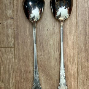 May include: Two silver serving utensils: a ladle and a salad fork. The ladle has a rounded bowl, while the fork has a slotted, petal-shaped head. Both have long, ornate handles with decorative detailing. The utensils are displayed on a wooden surface.