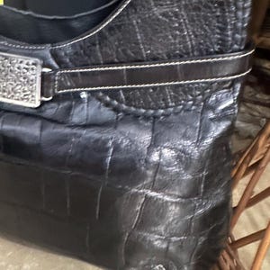 Puede incluir: Bolso de mano de cuero negro con un patr&oacute;n de cocodrilo texturizado. El bolso presenta una hebilla decorativa plateada y una correa marr&oacute;n oscuro con costuras blancas. La correa para el hombro tambi&eacute;n es negra con costuras blancas.