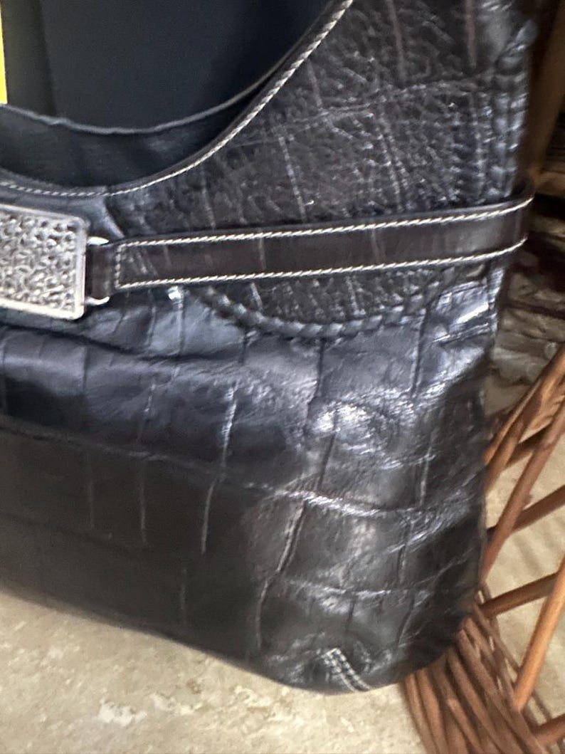 Puede incluir: Bolso de mano de cuero negro con un patr&oacute;n de cocodrilo texturizado. El bolso presenta una hebilla decorativa plateada y una correa marr&oacute;n oscuro con costuras blancas. La correa para el hombro tambi&eacute;n es negra con costuras blancas.