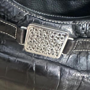 Puede incluir: Primer plano de un bolso de mano de cuero negro con un patr&oacute;n texturizado similar al de un cocodrilo. El bolso presenta una hebilla rectangular decorativa de color plateado con un intrincado dise&ntilde;o calado. Costuras blancas acent&uacute;an el cuero.