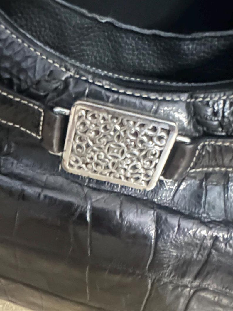 Puede incluir: Primer plano de un bolso de mano de cuero negro con un patr&oacute;n texturizado similar al de un cocodrilo. El bolso presenta una hebilla rectangular decorativa de color plateado con un intrincado dise&ntilde;o calado. Costuras blancas acent&uacute;an el cuero.