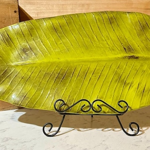 May include: A large, leaf-shaped serving platter in a vibrant yellow-green hue, with dark veining details. The platter is supported by a black, wrought-iron stand. This decorative piece adds a touch of nature-inspired elegance to any table setting.