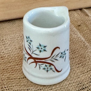 May include: A small, white ceramic creamer with a curved handle. The creamer is decorated with a brown floral vine design and small blue flowers. The creamer sits on a tan burlap surface.