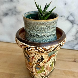May include: A small succulent plant in a blue and brown ceramic pot, nested inside a larger, decorative pot. The larger pot features a detailed, hand-painted design with various colors and patterns, including birds and floral elements. The pots are on a wooden surface.