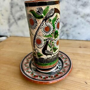 May include: A hand-painted ceramic vase with a matching saucer. The vase features a detailed design of birds, flowers, and foliage in black, green, and brown against a cream background. The saucer has a similar floral pattern with red accents.