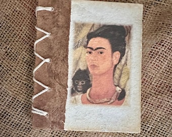 May include: A handmade journal with a brown textured cover and a white paper section featuring a portrait of Frida Kahlo. The journal is bound with white thread, and the background is a burlap texture.