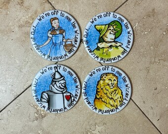 May include: Set of four round buttons featuring characters from The Wizard of Oz. Each button has a different character illustration: Dorothy, the Scarecrow, the Tin Man, and the Cowardly Lion. The text "We're off to see the Wizard" encircles each image.