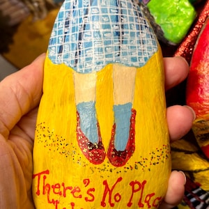 May include: Hand-painted rock featuring a yellow brick road design with red shoes and blue socks. The top of the rock has a blue and white checkered pattern. The text "There's No Place Like Home" is written in red.