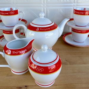 May include: A white porcelain tea set with red and gold accents. The set includes a teapot, sugar bowl, creamer, and four teacups with saucers. Each piece is decorated with the text "Beijing 2008" and Olympic rings.