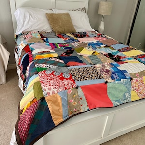 May include: A colorful patchwork quilt with a variety of fabric patterns and colors, including red, blue, and floral prints, covers a white bed. The quilt is on a white bed frame with white pillows and a tan pillow.