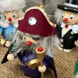 May include: A wooden pirate figurine wearing a maroon felt hat with a gold trim and a clear rhinestone, a gray faux fur beard, and an eye patch. The figure has a red nose, a purple body, a gold chain, and holds a gold staff with a red gem on top.