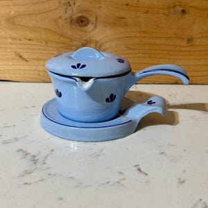 May include: A light blue enamelware sauce pot with a matching lid and a small handle. The pot sits on a matching saucer with a handle. Dark blue floral designs decorate the pot and lid. The pot is approximately 4 inches in diameter.