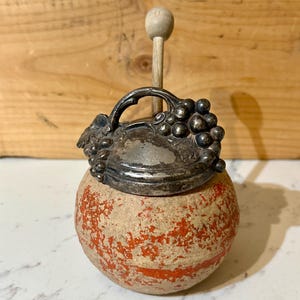 May include: A vintage, round container with a silver lid and a wooden handle. The lid is adorned with a grape cluster design. The container has a red and beige speckled pattern.