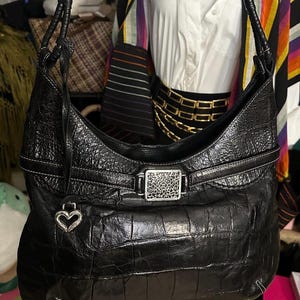 Puede incluir: Bolso de hombro de cuero negro con un patr&oacute;n de cocodrilo texturizado. El bolso presenta una hebilla plateada con un dise&ntilde;o decorativo y un dije en forma de coraz&oacute;n. Costuras blancas acent&uacute;an los bordes.