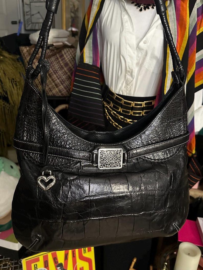 Puede incluir: Bolso de hombro de cuero negro con un patr&oacute;n de cocodrilo texturizado. El bolso presenta una hebilla plateada con un dise&ntilde;o decorativo y un dije en forma de coraz&oacute;n. Costuras blancas acent&uacute;an los bordes.