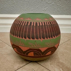 May include: A spherical ceramic vase featuring a matte finish with horizontal bands of sage green, terracotta, and dark brown. The surface is decorated with carved geometric patterns, including vertical lines, stylized grass motifs, and semi-circles.
