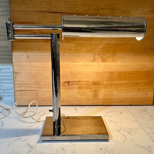 May include: A chrome desk lamp with a cylindrical shade and a long, adjustable arm. The lamp has a simple, modern design and is in excellent condition.