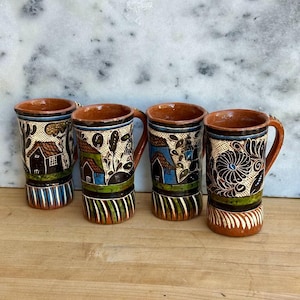 May include: Four handcrafted ceramic mugs with intricate designs. Each mug features a brown handle and rim, with colorful patterns of houses, trees, and floral motifs on a cream background. The bases have vertical stripes in green, orange, and white.