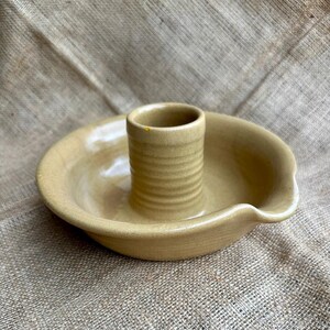 May include: A beige ceramic candle holder featuring a wide, shallow circular base with a raised, ribbed cylindrical center. The piece has a smooth, glazed finish and a small, curved indentation along the outer rim of the bowl-shaped base.