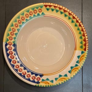 May include: A large, off-white ceramic bowl with a colorful geometric design around the rim. The rim features alternating patterns of red, blue, yellow, and green triangles and diamond shapes. The bowl has a wide, shallow shape.