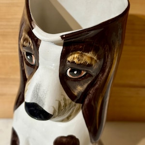 May include: A ceramic mug shaped like a brown and white basset hound dog. The mug has a white interior and brown and white painted details.