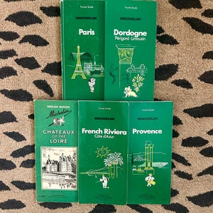 May include: A collection of five green Michelin tourist guides featuring white text and line art illustrations. The covers include titles for Paris, Dordogne, Chateaux of the Loire, French Riviera, and Provence, each displaying iconic regional landmarks.