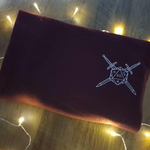 May include: A burgundy pouch featuring a white graphic of a twenty-sided die and two crossed swords. The rectangular pouch is made of a soft material. The background has warm fairy lights.