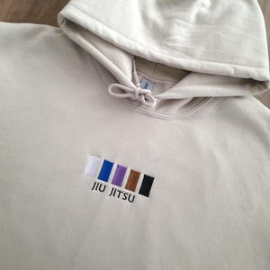 May include: A beige hoodie with a drawstring hood and a small embroidered design of coloured squares and the text "JIU JITSU".