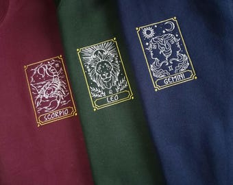 Puede incluir: Tres sudaderas con signos del zodíaco bordados. La sudadera granate tiene un escorpión, la sudadera verde tiene un león y la sudadera azul marino tiene gemelos.