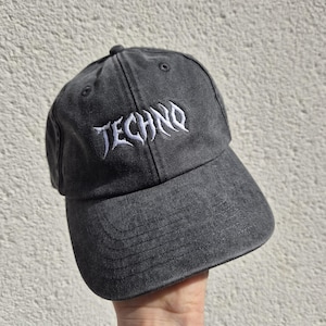 Könnte beinhalten: Eine anthrazitfarbene Baseballkappe mit verwaschenem Vintage-Finish. Auf der Vorderseite ist das Wort TECHNO in einer stilisierten, scharfen weißen Schrift eingestickt. Die Kappe verfügt über einen gebogenen Schirm und gestickte Ösen zur Belüftung.