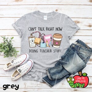 May include: A grey t-shirt with the text "Can't talk right now doing teacher stuff" and a graphic of a coffee cup, notebook, pencils, and other teacher supplies. The shirt is on a wooden surface with a pair of white trainers and blue jeans.