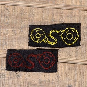 May include: Two black fabric patches with yellow and red embroidered designs of handcuffs. The handcuffs are connected by a chain.