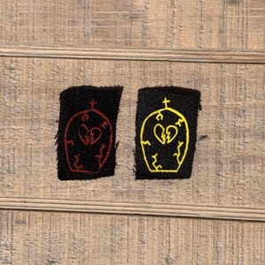 May include: Two black fabric patches with a yellow and red embroidered design of a broken heart inside a tombstone with a cross on top.
