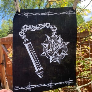 Pode incluir: Bandana preta com um desenho branco de uma clava com espinhos e uma rosa. A bandana tem uma borda de arame farpado branco.