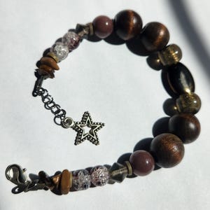Puede incluir: Una pulsera de cuentas con una mezcla de cuentas de madera, vidrio y piedra en tonos marrón, morado y transparente. La pulsera incluye un dije de estrella plateada y un cierre de mosquetón. Las cuentas varían en tamaño y forma.