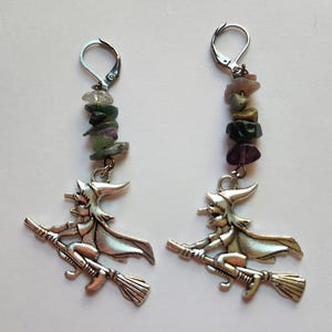 May include: A pair of silver-toned earrings featuring a witch flying on a broomstick charm suspended from a vertical stack of small, multi-colored tumbled gemstone chips and a silver-toned lever-back ear wire closure for secure attachment.