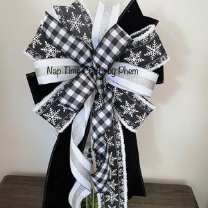Pode incluir: Um laço decorativo com fitas xadrez em preto e branco e com padrões de flocos de neve, acentuado com fitas prateadas e brancas. O laço está preso a uma lanterna preta e branca. O texto no laço diz "Nap Time Crafts by Phem."