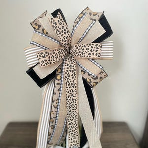 May include: A decorative bow featuring a mix of patterned ribbons. The bow includes ribbons with leopard print, black and white gingham, and striped designs. The bow is set on a white base, with the text "Nap Time Crafts by Phem" visible at the bottom.