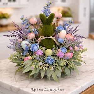 May include: An Easter centerpiece featuring a moss-covered bunny surrounded by pastel-colored flowers and eggs. The arrangement includes blue, pink, and purple blooms, with green foliage. The base is a white container, creating a festive spring display.