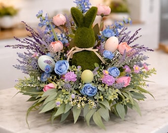 Peut inclure: Une composition florale de Pâques avec un lapin recouvert de mousse entouré de fleurs et d'œufs aux couleurs pastel. L'arrangement comprend des fleurs bleues, roses et violettes, avec un feuillage vert. La base est un contenant blanc.