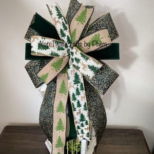 Pode incluir: Um laço de Natal decorativo com várias fitas verdes, douradas e bege. O laço apresenta um centro de veludo verde escuro e fitas com padrões de árvores de Natal e veados. O texto "Nap Time Crafts by Phem" é visível numa das fitas.