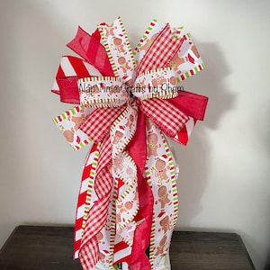 Pode incluir: Um laço de Natal festivo com fitas listradas em vermelho e branco, xadrez e com estampa de boneco de gengibre. O laço está em cima de uma lanterna branca, criando uma decoração de férias. O texto "Nap Time Crafts by Phem" é visível.