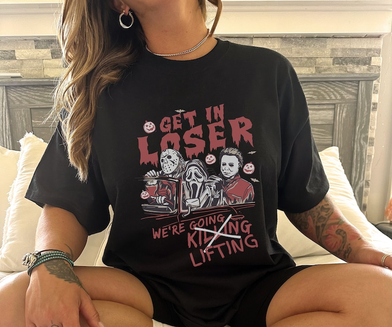 May include: Black t-shirt featuring a graphic of three horror movie characters in a car. The text on the shirt reads "Get in Loser, We're Going Killing Lifting" with the word "Killing" crossed out.