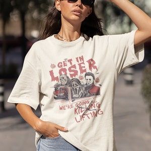 May include: A white t-shirt featuring a graphic of three horror movie characters in a car. The text on the shirt reads "Get in Loser, We're Going Killing Lifting".