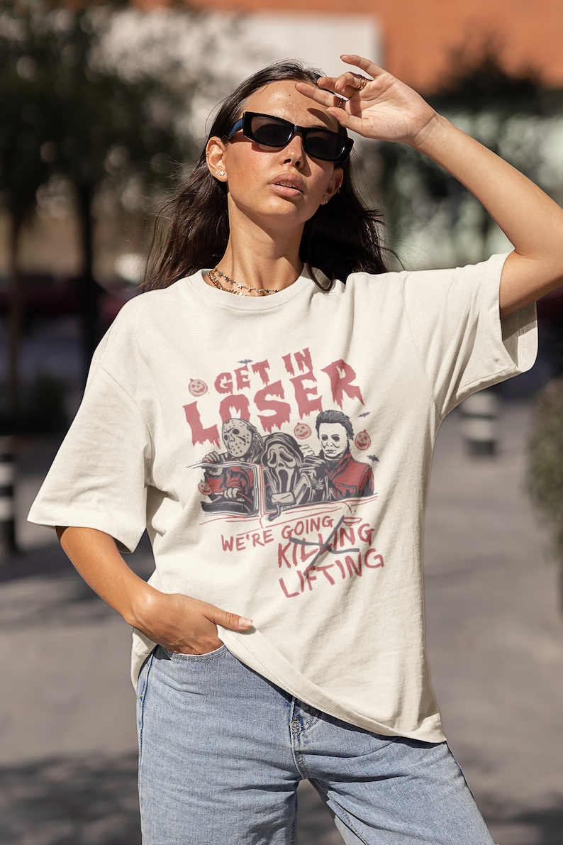 May include: A white t-shirt featuring a graphic of three horror movie characters in a car. The text on the shirt reads "Get in Loser, We're Going Killing Lifting".