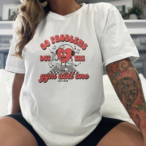Puede incluir: Camiseta blanca con un gr&aacute;fico de coraz&oacute;n rojo y el texto "99 problemas pero el gimnasio no es uno" en una fuente retro. El coraz&oacute;n lleva auriculares y sostiene pesas. El texto "L&E Crew" est&aacute; debajo del gr&aacute;fico.