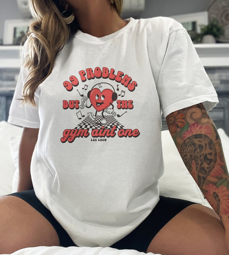 Puede incluir: Camiseta blanca con un gr&aacute;fico de coraz&oacute;n rojo y el texto "99 problemas pero el gimnasio no es uno" en una fuente retro. El coraz&oacute;n lleva auriculares y sostiene pesas. El texto "L&E Crew" est&aacute; debajo del gr&aacute;fico.