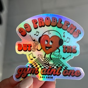 Puede incluir: Una pegatina hologr&aacute;fica con un coraz&oacute;n rojo que lleva auriculares y sostiene pesas. La pegatina dice "99 problemas pero el gimnasio no es uno" y "L&E Crew" en la esquina inferior derecha.